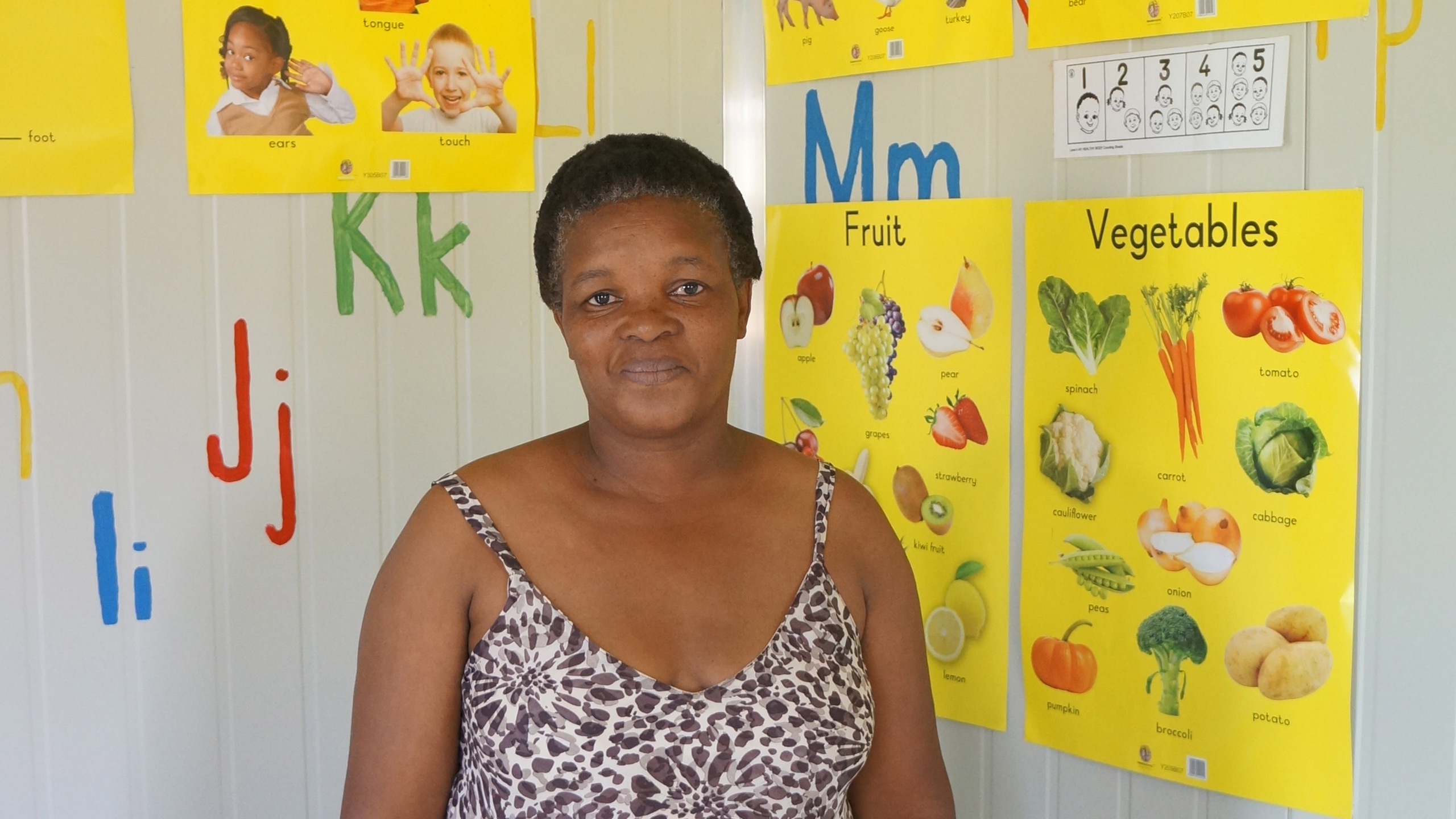 KITA-Leiterin Gladys vor bunten Unterrichtspostern – die Kinder sind begeistert vom neuen Schulalltag und kommen am liebsten täglich.