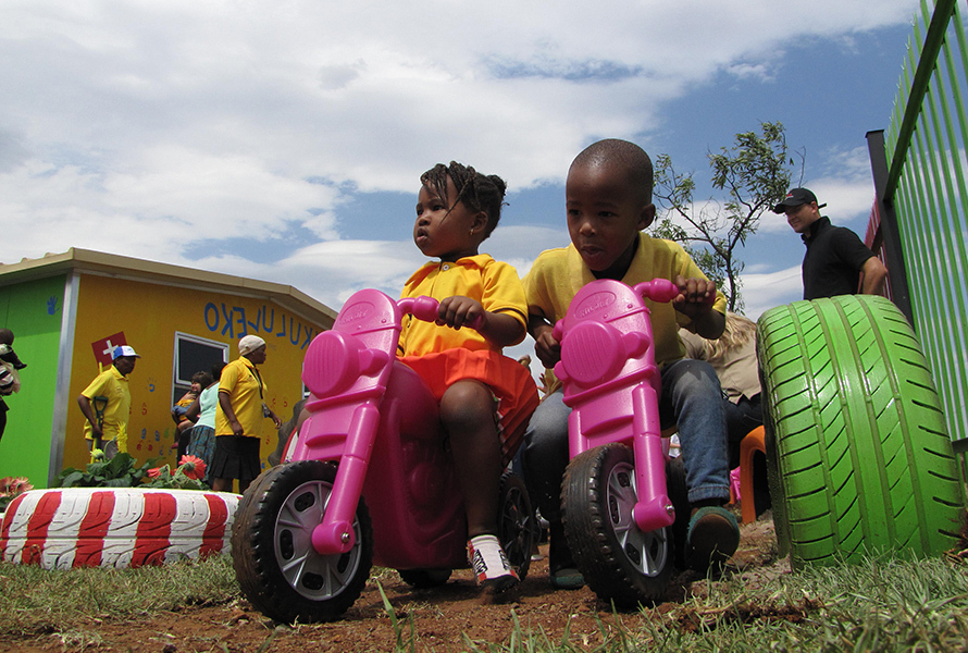 Ein Junge und ein Mädchen der KITA Nkululeko sitzen lachend auf pinken Plastikmotorrädern – voller Energie und Lebensfreude.