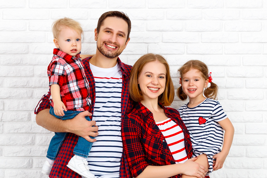 La famiglia al centro Una famiglia con due bambini è riunita davanti a un muro di mattoni bianchi – un momento di vicinanza, come PRO LIFE desidera promuovere per tutte le famiglie.