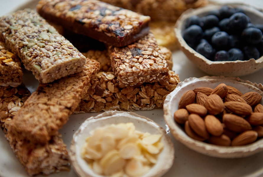 Müesliriegel mit Nüssen und Beeren Verschiedene hausgemachte Müesliriegel auf einem Teller, daneben kleine Schälchen mit Blaubeeren, Mandeln und Mandelplättchen – der perfekte Energiespender.