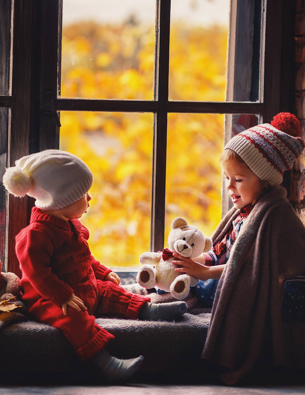 Moment entre frère et sœur à la fenêtre Une fille et un garçon en habits d’hiver sont assis confortablement sur le rebord de la fenêtre, la fille tient un ours en peluche – un instant plein de chaleur et de sécurité.