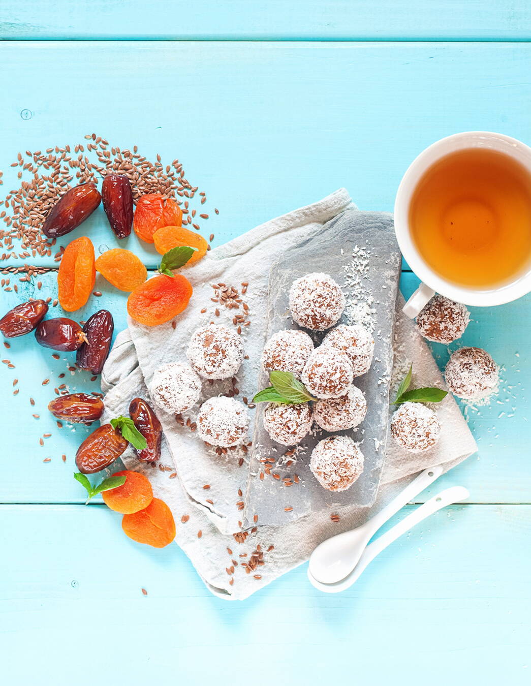 Deliziose palline per l’allattamento con ingredienti naturali e tè Powerballs a base di datteri, albicocche e semi di lino su un tovagliolo, circondati da una tazza di tè e ingredienti freschi – lo spuntino ideale per ricaricarsi di energia durante il post parto e l’allattamento.