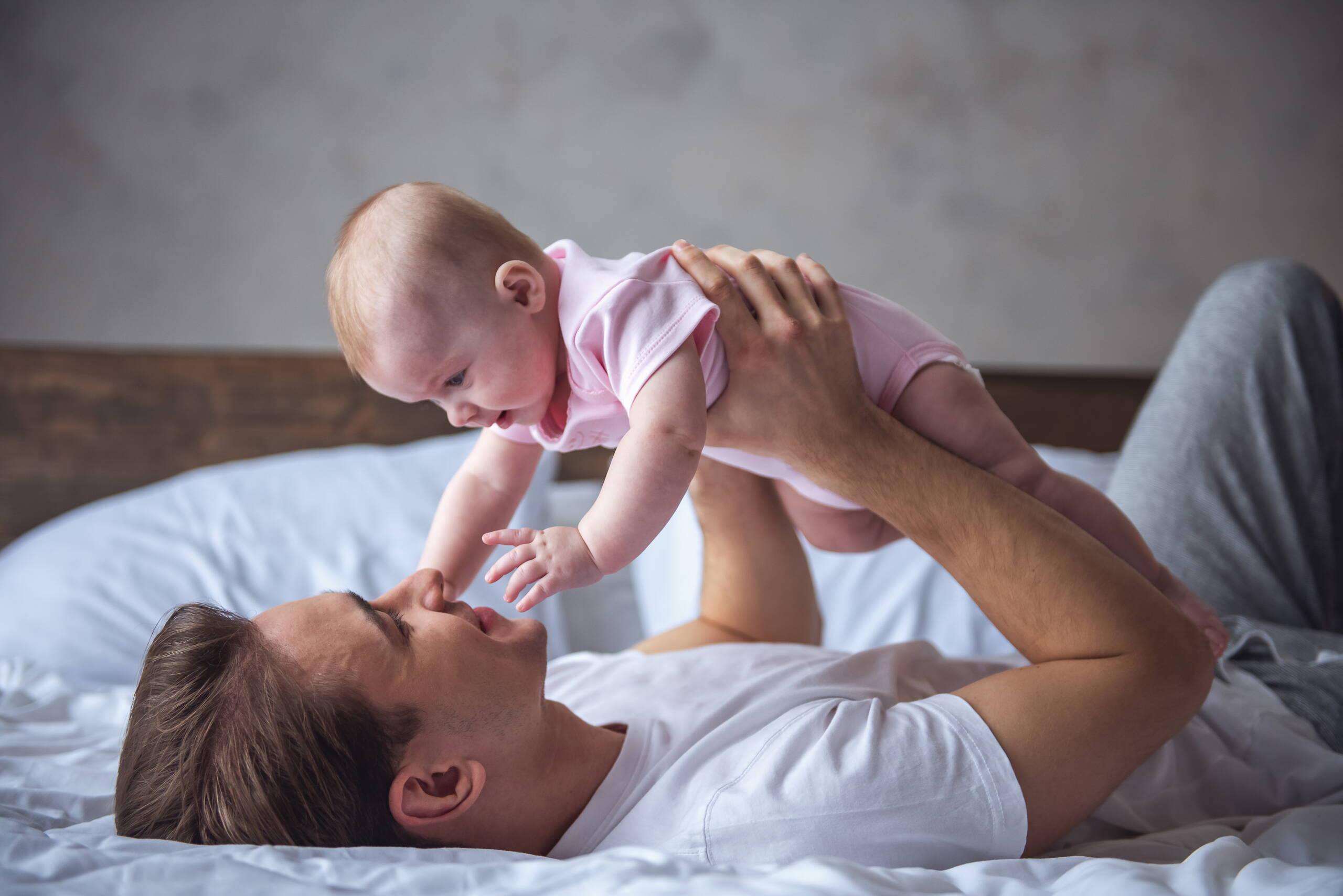 Père et enfant dans un moment de confiance Un jeune père est allongé sur le dos, détendu, tenant tendrement son enfant au-dessus de lui – une image de sécurité et de proximité.
