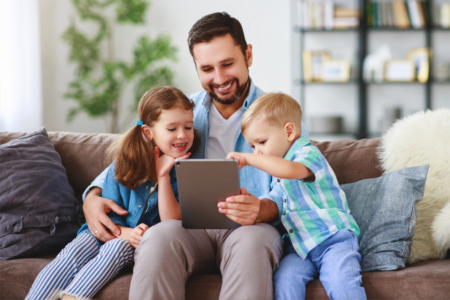 Gemeinsame Momente Ein Vater sitzt mit seinen zwei Kindern auf dem Sofa und schaut auf ein Tablet – ein Symbol für Nähe, Unterstützung und informierte Entscheidungen.