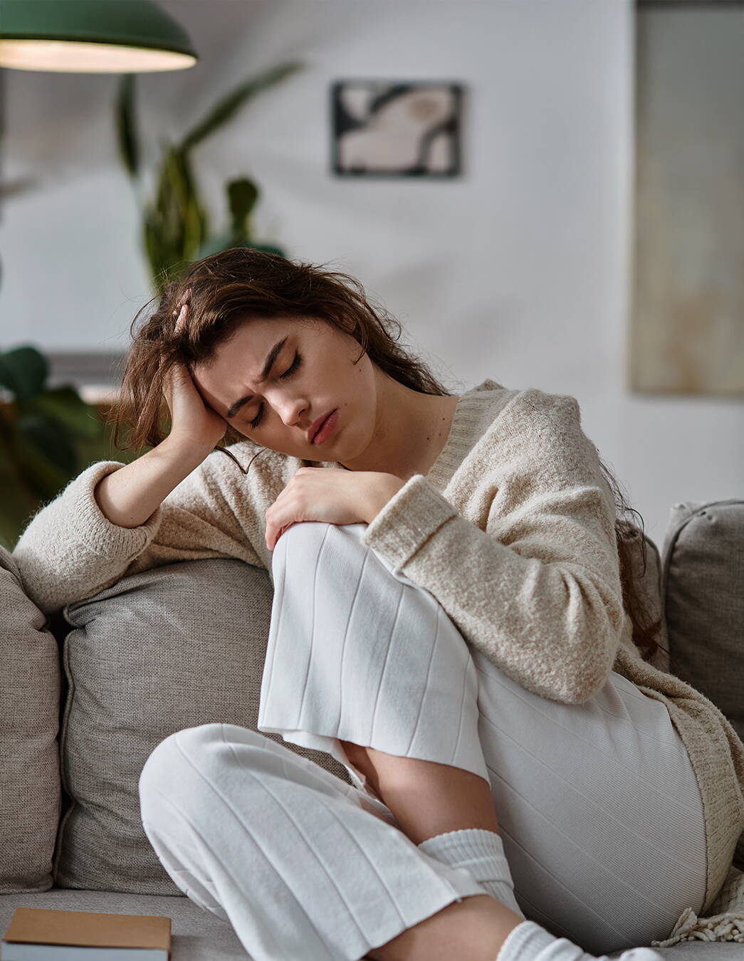 Fatigue pendant la grossesse – femme enceinte fatiguée assise sur le canapé Femme enceinte assise sur un canapé, fatiguée, la tête appuyée sur la main – fatigue typique pendant la grossesse au quotidien.