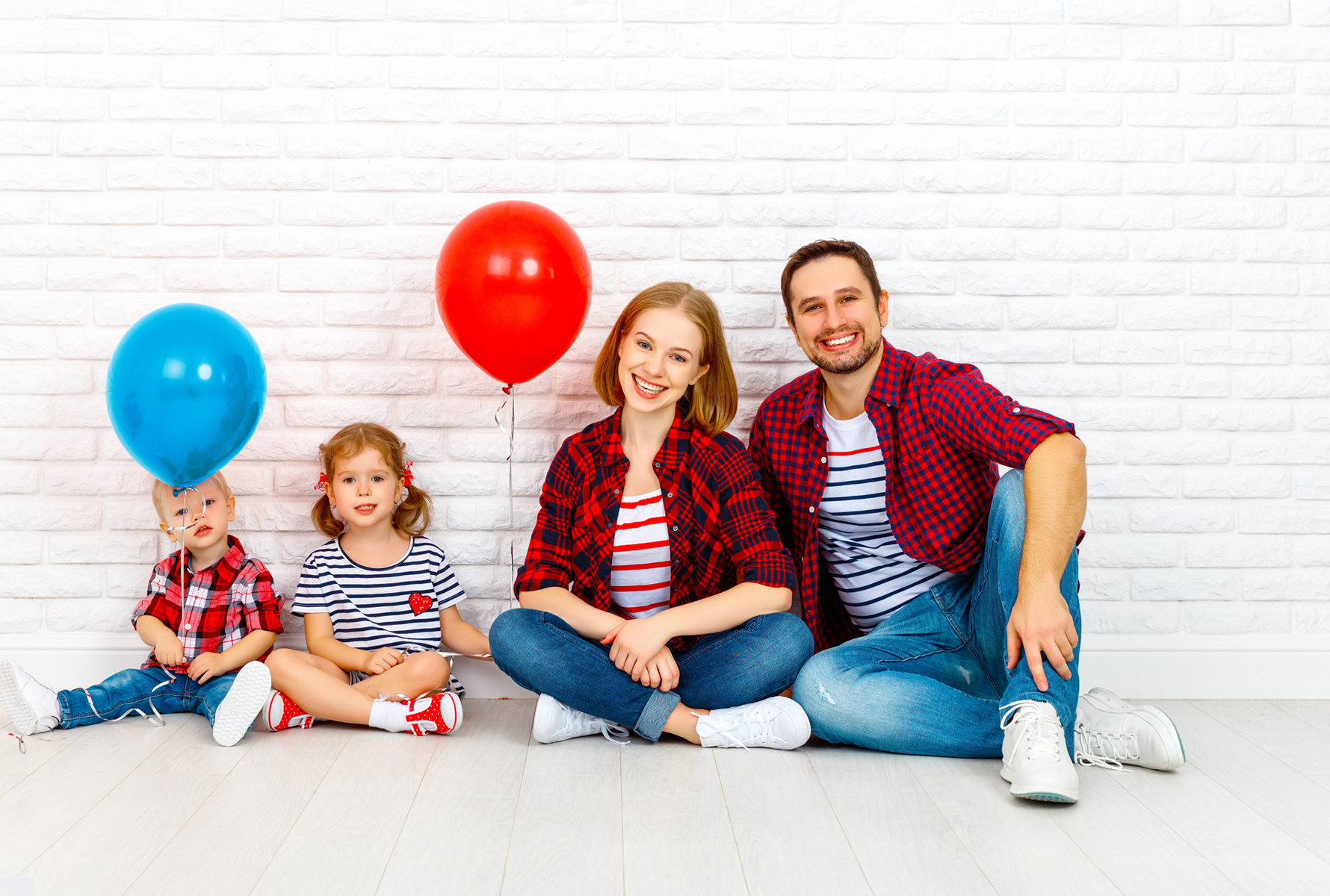 Famille avec ballons devant un mur en briques blanches La même famille est assise détendue devant un mur en briques blanches, les enfants tiennent un ballon rouge et un ballon bleu – un moment plein de légèreté.