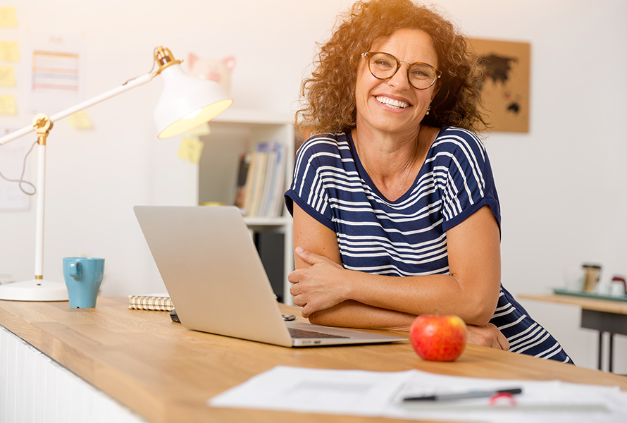 Télétravail à domicile Une personne est assise à un bureau avec un ordinateur portable et sourit à la caméra. Sur le bureau se trouvent une lampe, une pomme et des papiers. En arrière-plan, on voit des étagères avec des livres et des notes au mur.