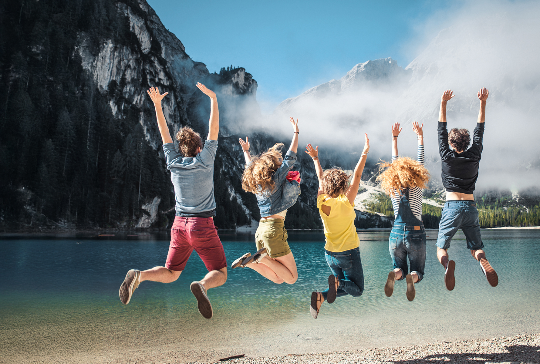 Loisirs avec vue Un groupe de jeunes saute en l’air au bord d’un lac de montagne – une image de liberté, de joie de vivre et des avantages loisirs pour les membres PRO LIFE.
