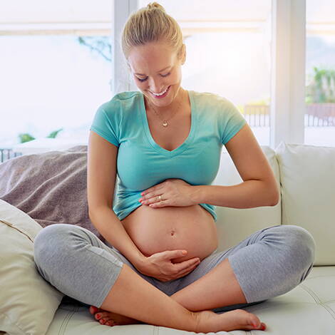 Gelassen in die Geburt Eine strahlende Schwangere sitzt im Schneidersitz auf dem Sofa und hält liebevoll ihren Bauch – ein Moment der Vorfreude und inneren Ruhe kurz vor dem grossen Ereignis.