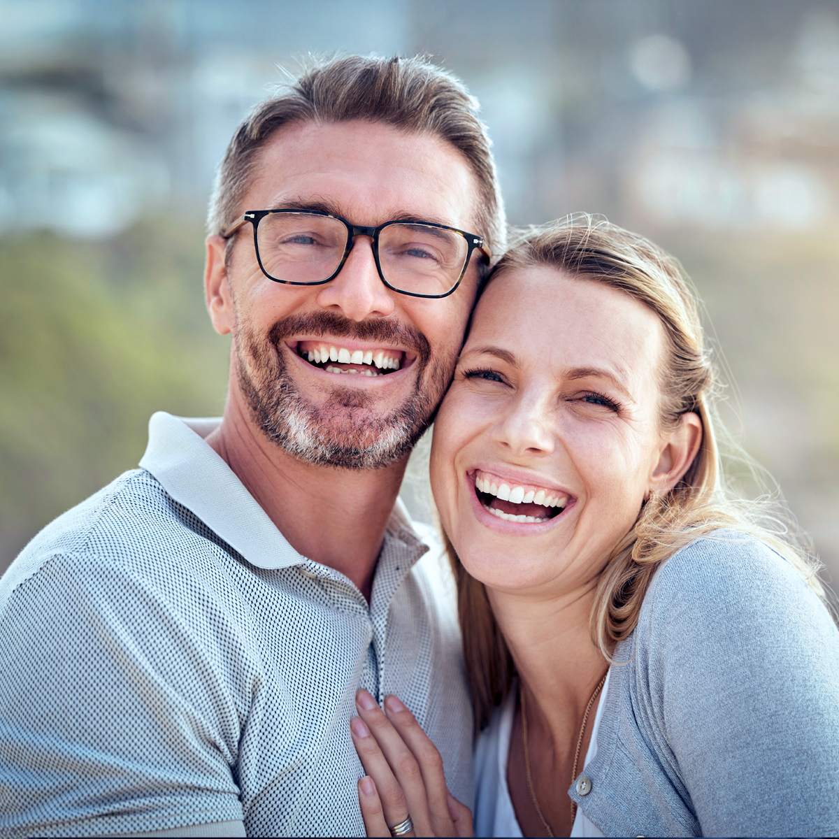 Ensemble en chemin Deux personnes sont côte à côte et sourient à la caméra. La personne à gauche porte des lunettes et une chemise claire, celle à droite un haut clair. L’arrière-plan flou montre un décor extérieur avec des bâtiments et de la verdure.