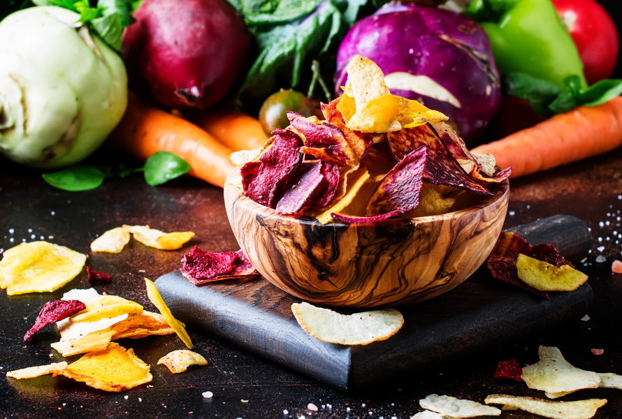 Knusprige Gemüsechips aus frischem Gemüse Holzschüssel mit bunten, selbstgemachten Gemüsechips aus Randen, Tomaten, Kohlrabi und Rüebli, vor frischem Gemüse auf Holztisch – kalorienarm und knackig.
