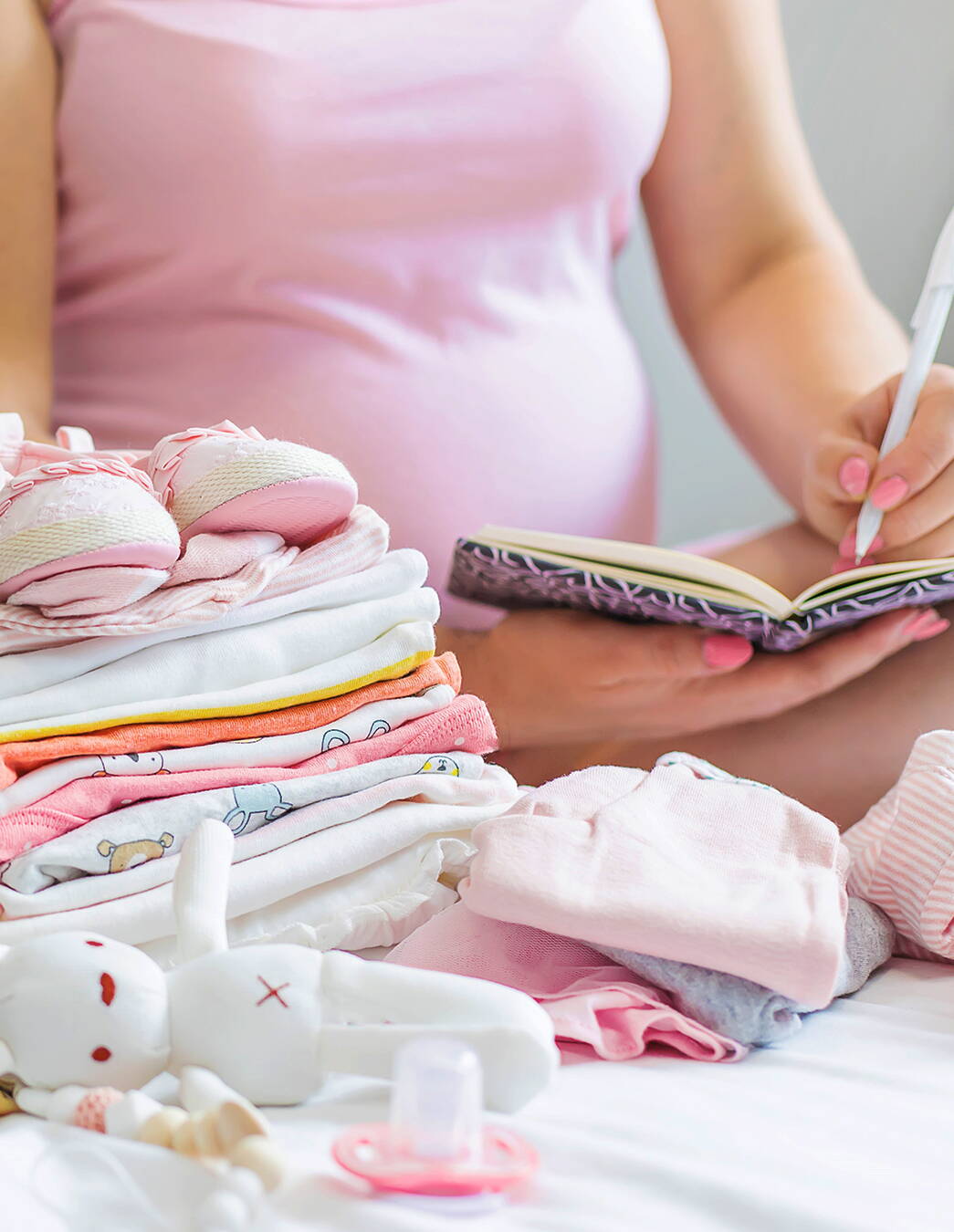 Eine schwangere Frau macht Notizen in ein Buch, im Vordergrund liegt liebevoll gefaltete Babykleidung – Erinnerungen für die Zukunft entstehen.