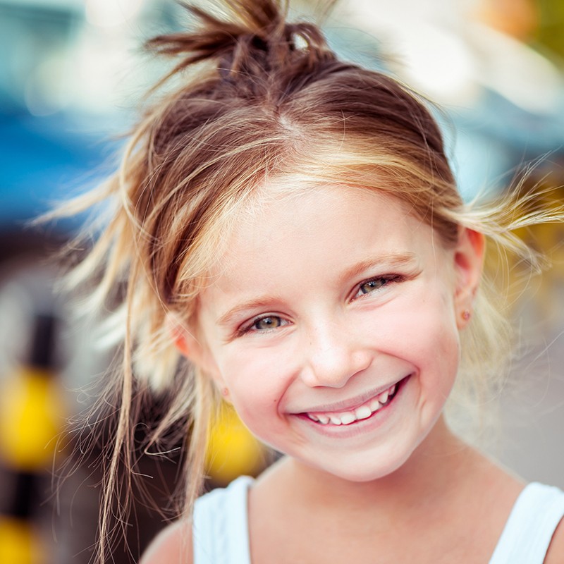 Un enfant aux cheveux en bataille et à un chignon lâche porte un haut sans manches. L’arrière-plan est flou, avec des éléments jaunes et noirs suggérant une rue ou un parking.