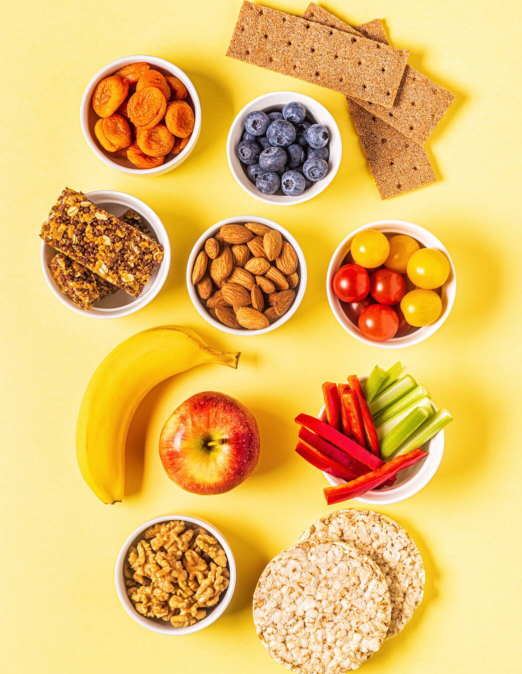 Collations saines contre les fringales pendant la grossesse Sur l’image, vous voyez des petits bols colorés contenant des abricots, des myrtilles, des barres de céréales, des amandes, des tomates cerises et des bâtonnets de légumes – le tout accompagné de pain croustillant complet, de galettes de riz et de fruits frais. Parfait pour combler une petite envie entre deux repas!