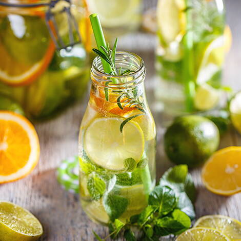 Aromatisiertes Wasser – frisch und gesund Flaschen mit Infused Water umgeben von frischen Zutaten wie Rosmarin, Melisse, Orangen, Zitronen und Limetten – lecker, kalorienarm und durstlöschend.