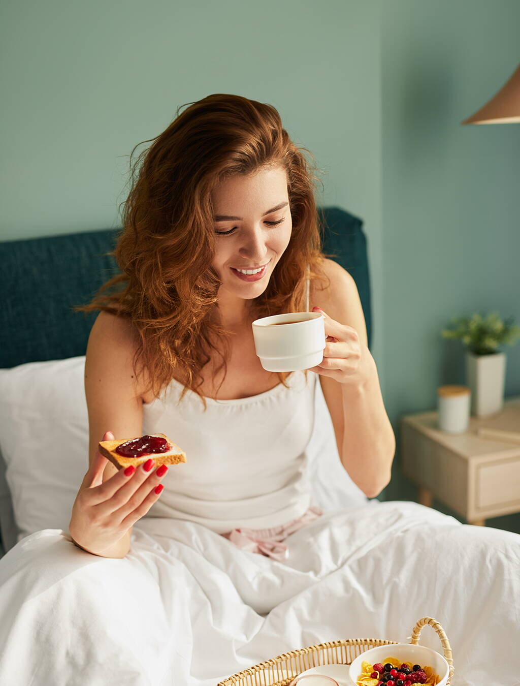 Eine Frau sitzt im Bett, hält eine Tasse in der einen und eine Scheibe Toast mit Konfitüre in der anderen Hand – ein Moment der Ruhe trotz Morgenübelkeit.