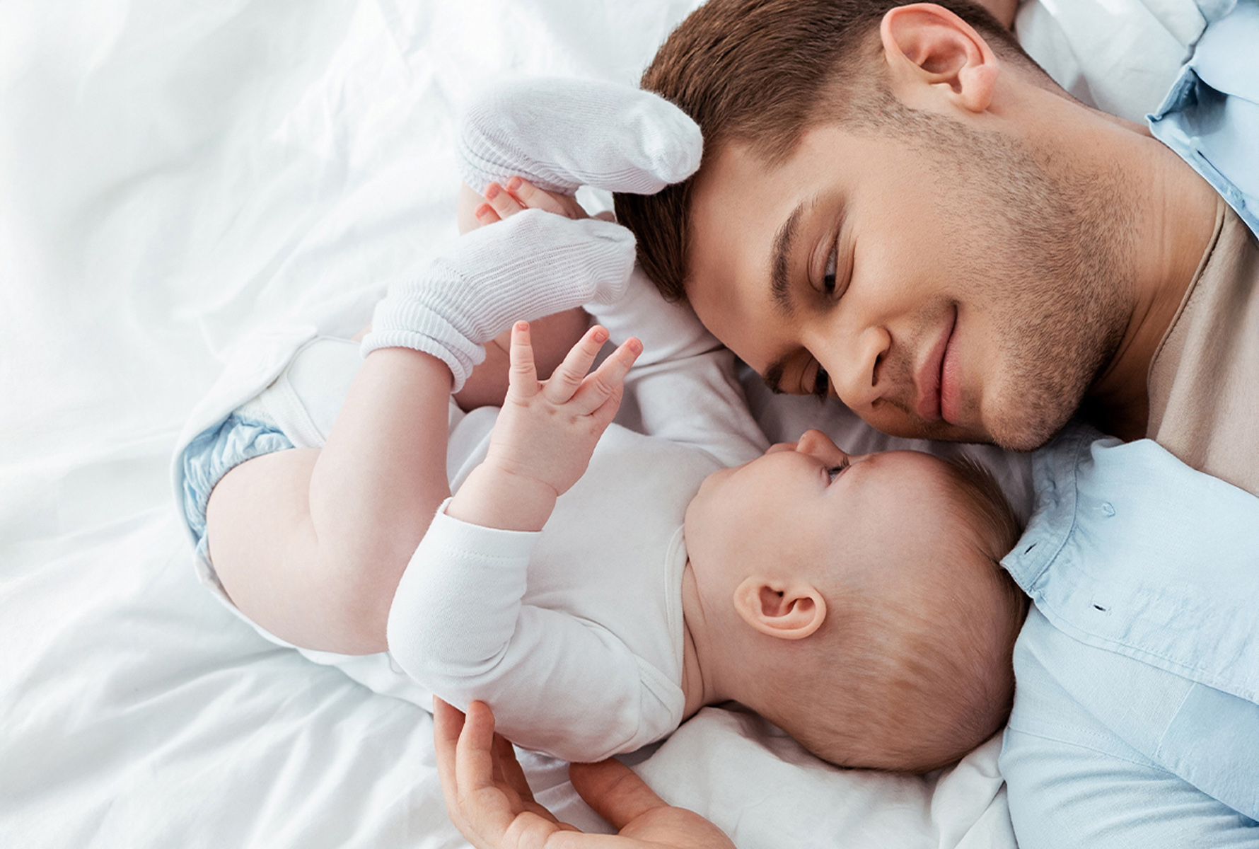 Il nostro impegno Un padre sdraiato a faccia a faccia con il suo bambino sul letto – un momento intimo che mostra ciò per cui PRO LIFE si impegna: vicinanza, famiglia, umanità.