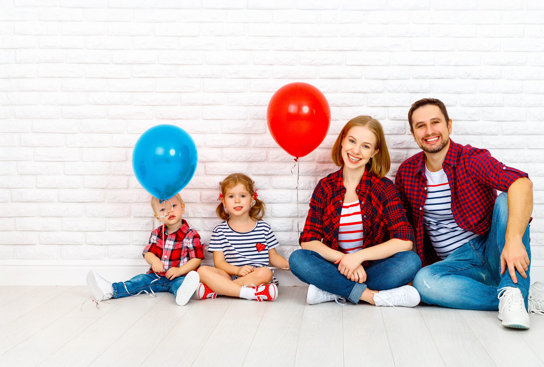 Die gleiche Familie sitzt entspannt vor einer weissen Backsteinmauer, die Kinder halten einen roten und blauen Ballon – ein Moment voller Leichtigkeit.