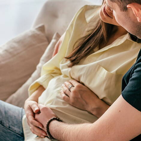 Paar versucht die Babybewegungen zu spüren Ein Paar sitzt entspannt auf dem Sofa, beide legen liebevoll die Hände auf den Bauch der Schwangeren – ein besonderer Moment der Verbundenheit.
