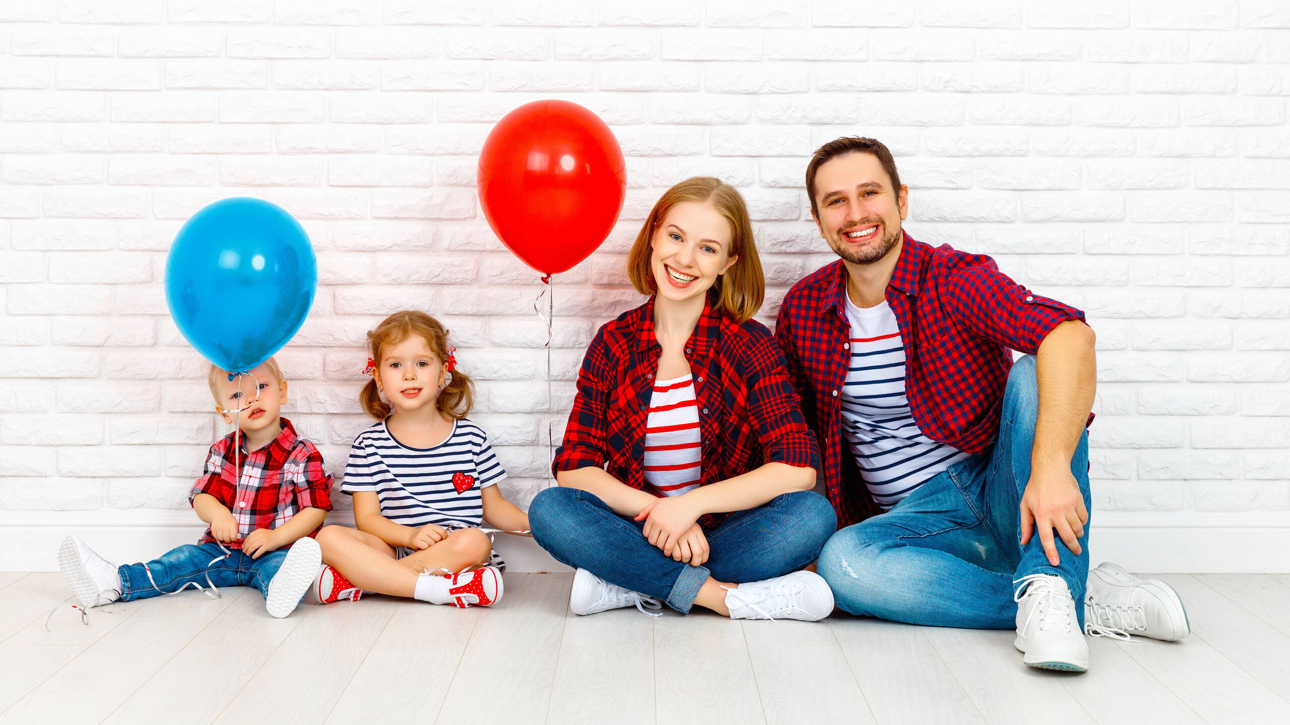 Di più per la vita – con PRO LIFE Una famiglia di quattro persone è seduta rilassata davanti a un muro chiaro in mattoni. I bambini tengono palloncini colorati – un simbolo di leggerezza e gioia di vivere con PRO LIFE.