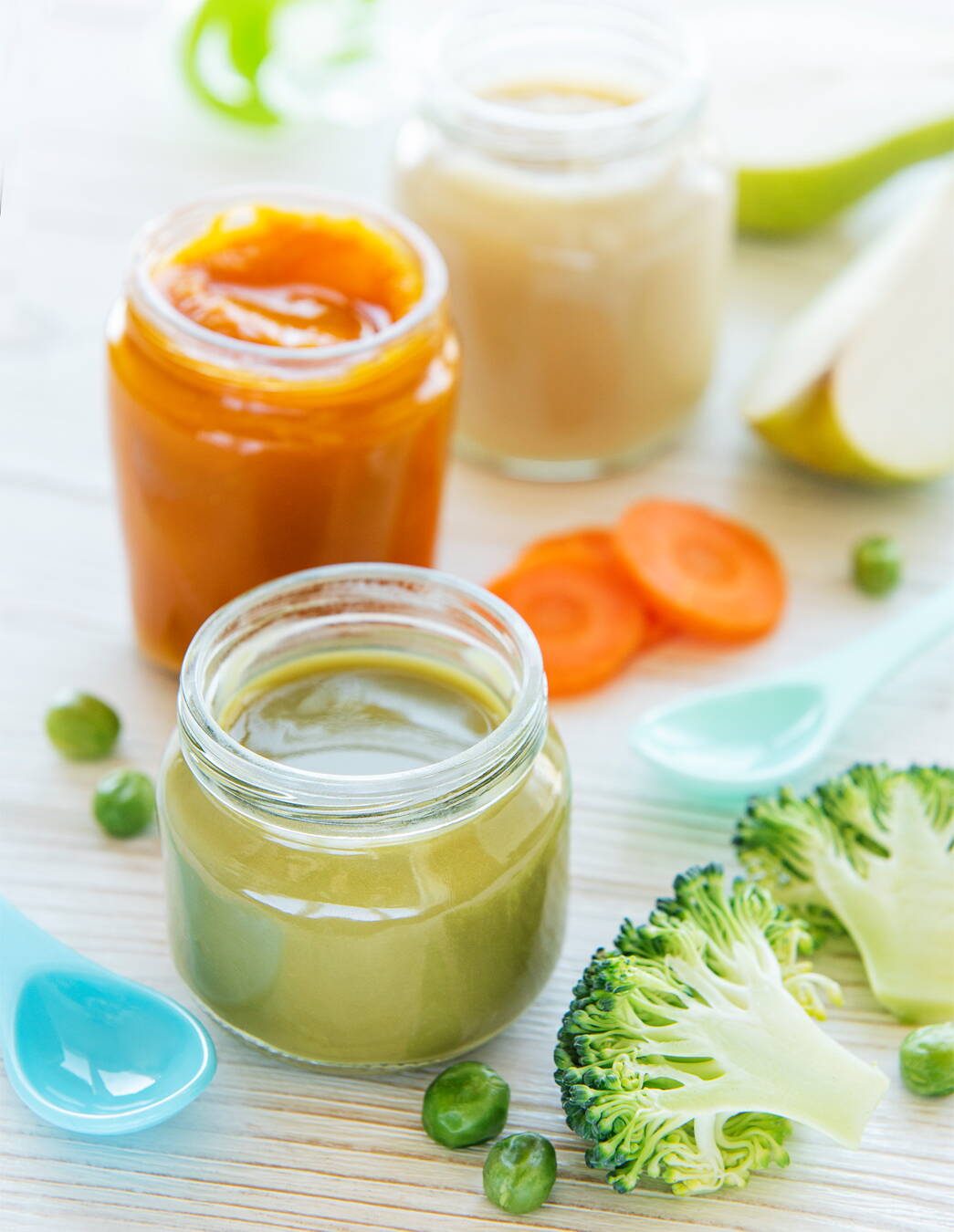Tre vasetti di pappa fatta in casa sono disposti su un tavolo di legno chiaro, accanto a cucchiaini per bebè, broccoli, piselli, carote e una mela – tutto è pronto per le prime cucchiaiate di divertimento

