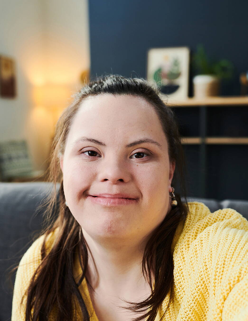 Porträt einer jungen Person mit Down-Syndrom Porträtaufnahme einer jungen Person mit Down-Syndrom in einem Innenraum. Die Person sitzt vor einem ruhigen Hintergrund und blickt in die Kamera. Warme Farben und eine angenehme Lichtstimmung schaffen eine freundliche Atmosphäre.