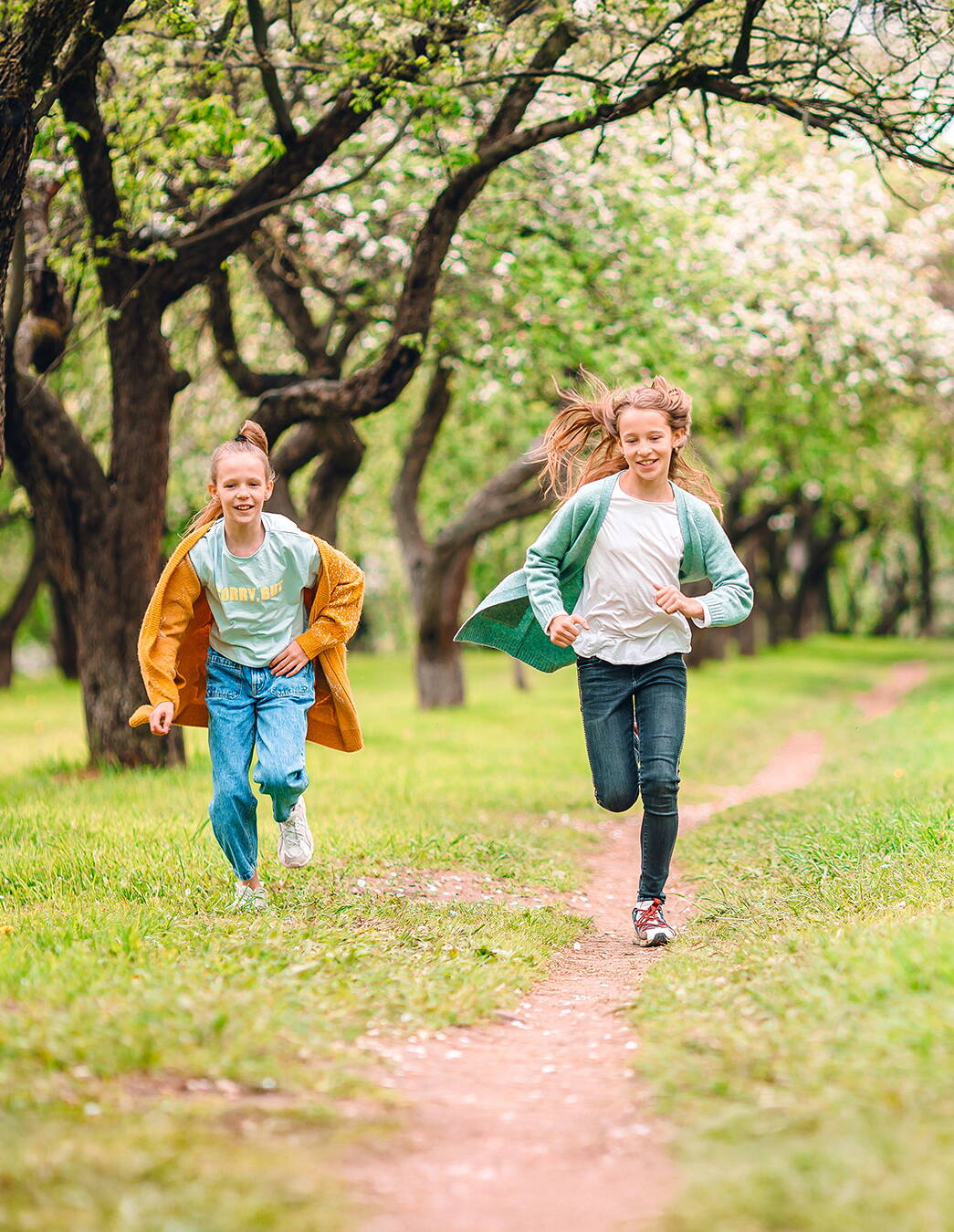 Deux enfants courent sur un chemin herbeux à travers un verger en pleine floraison, accompagnés d’un petit chien blanc. Les arbres sont en pleine floraison et créent une atmosphère printanière et naturelle.

