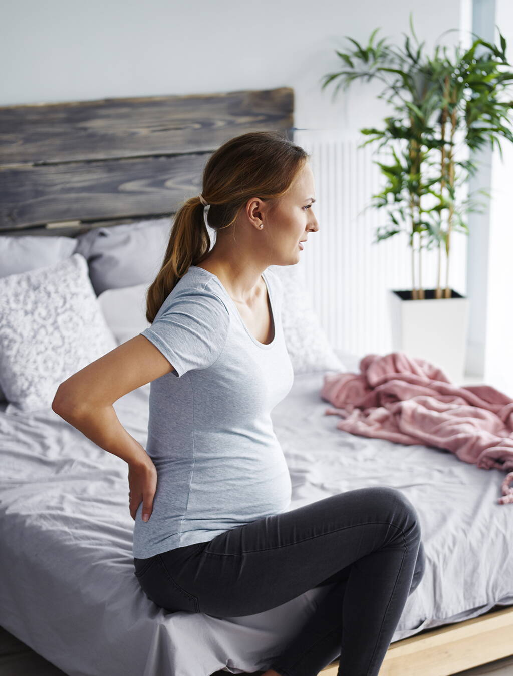 Schwangere Frau sitzt auf der Bettkante und hält sich den schmerzenden Rücken – ein typisches Problem, das viele werdende Mütter beschäftigt.