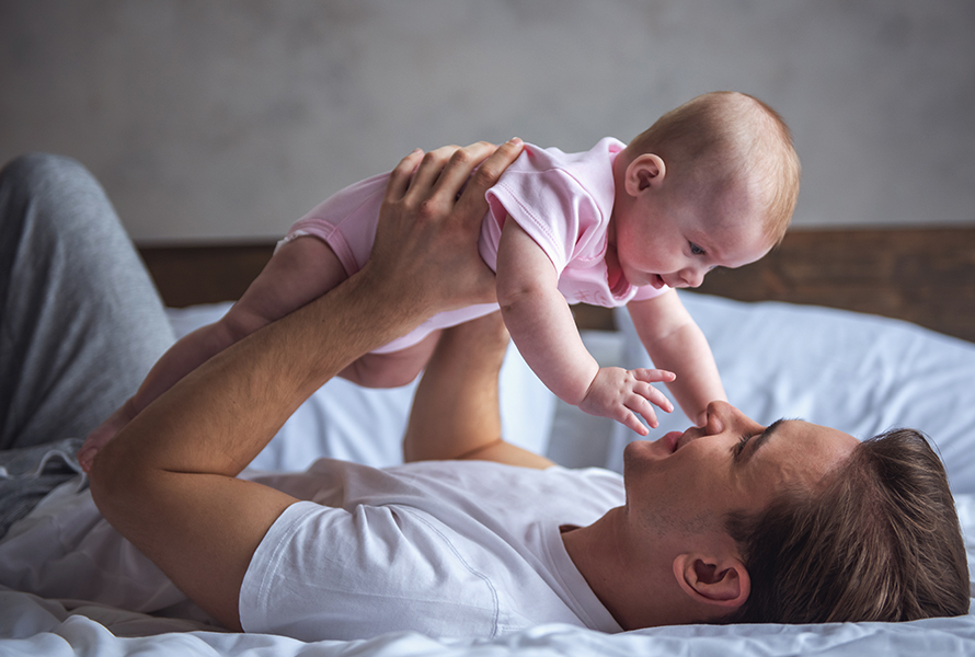 Ein Vater liegt entspannt auf dem Bett und hält sein Baby liebevoll über sich in die Höhe. Die beiden teilen einen innigen Blickkontakt – ein Moment voller Vertrauen, Freude und Geborgenheit.