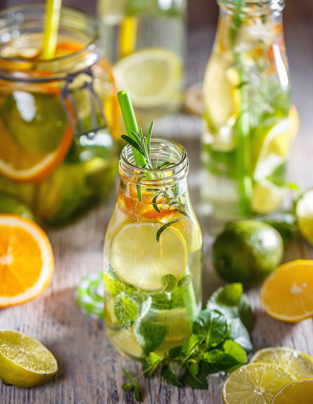 Aromatisiertes Wasser – frisch und gesund Flaschen mit Infused Water umgeben von frischen Zutaten wie Rosmarin, Melisse, Orangen, Zitronen und Limetten – lecker, kalorienarm und durstlöschend.
