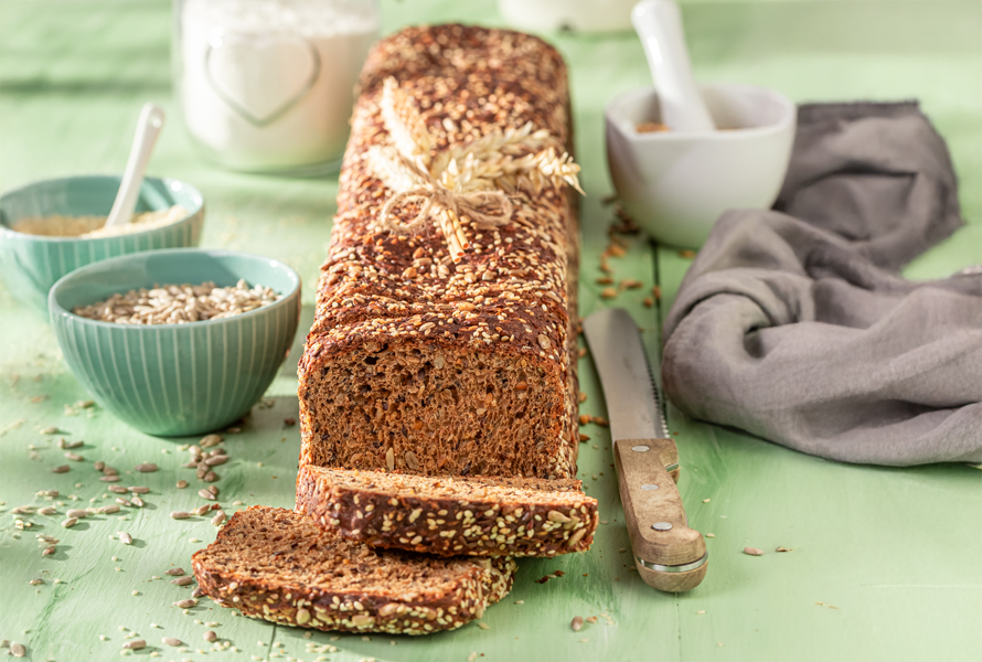 Frisch gebackenes Vollkornbrot Kastenvollkornbrot mit zwei angeschnittenen Scheiben auf grünem Holztisch, daneben ein Brotmesser, Schüsseln mit Körnern und ein Glas – sättigend und gesund.