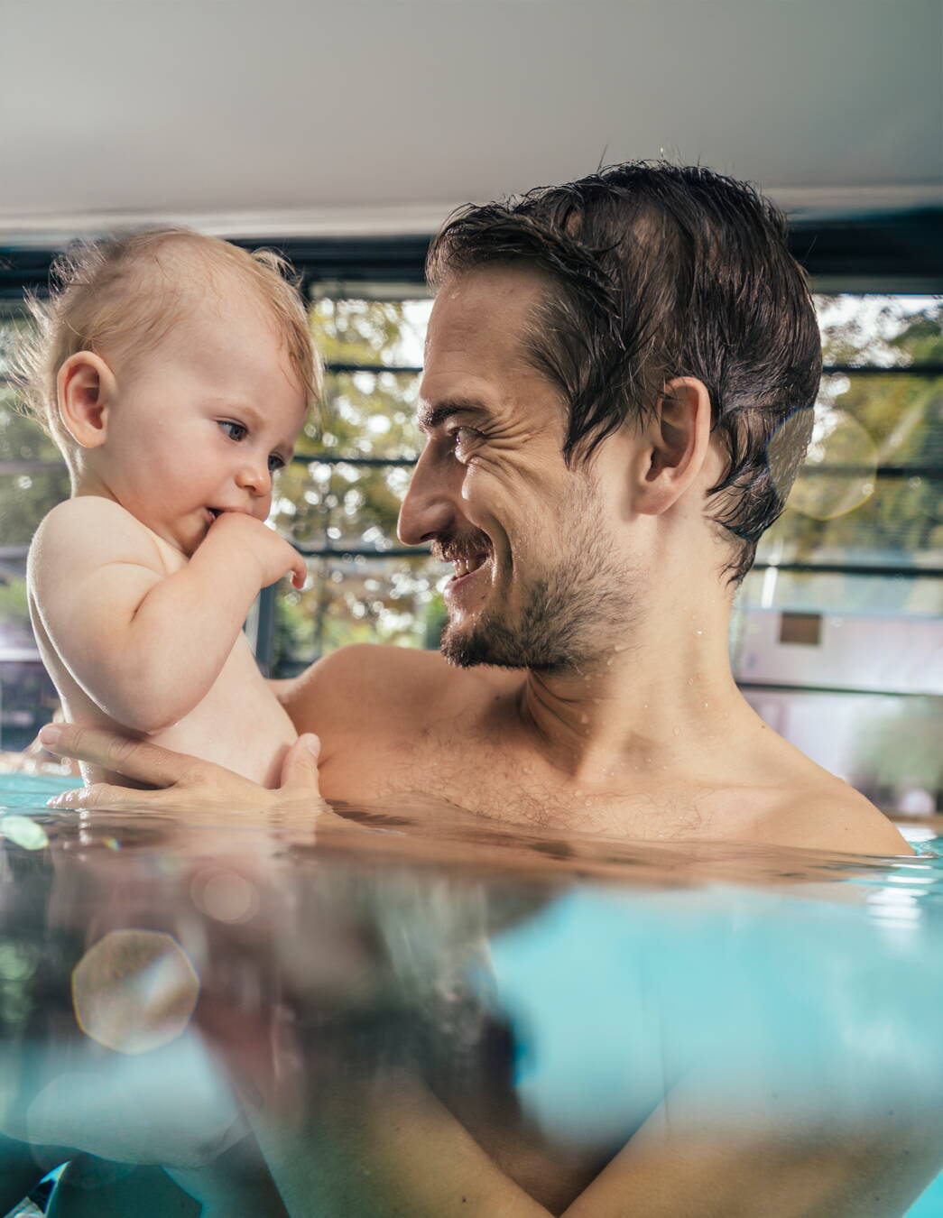 Ein Mann hält sein Kleinkind auf dem Arm, beide lachen im warmen Wasser eines Hallenbads – Nähe und Vertrauen beim Babyschwimmen.