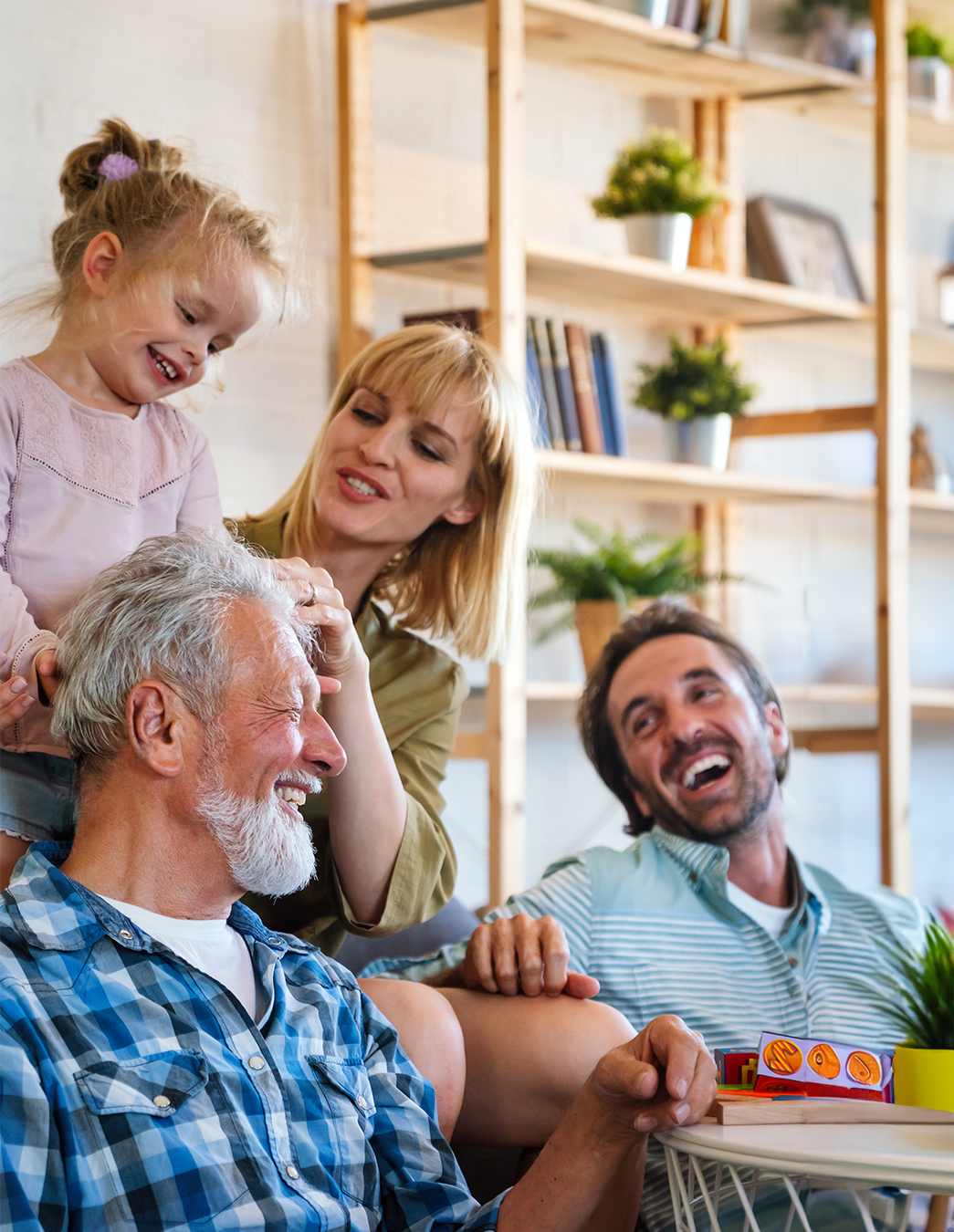 Gemeinsam lachen – Generationen im Austausch In einem warmen, wohnlichen Raum mit Bücherregalen und Pflanzen teilen vier Menschen einen fröhlichen Moment. Ein älterer Mann sitzt im Vordergrund, während eine Frau und ein Kind liebevoll mit ihm interagieren. Eine weitere erwachsene Person beobachtet die Szene lächelnd. Ein Bild voller Nähe und Lebensfreude.