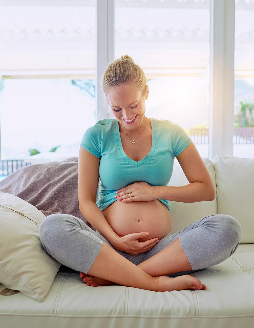 Une femme enceinte souriante est assise en tailleur sur le canapé et tient tendrement son ventre. Ce moment symbolise le soutien offert par les prestations de la caisse-maladie pendant la grossesse en Suisse.