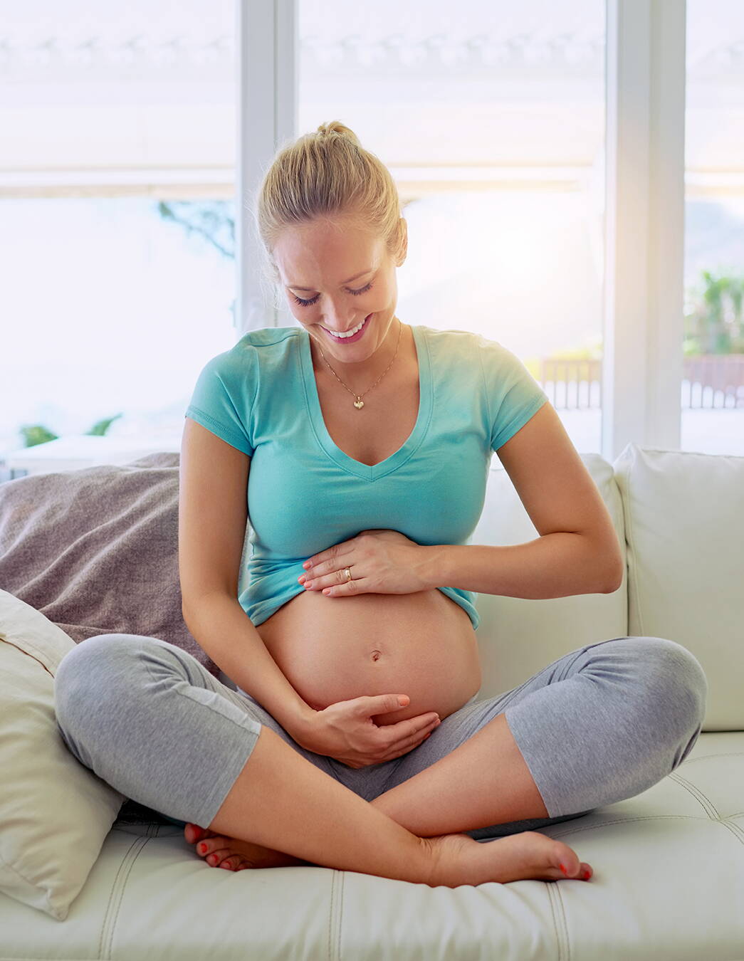Gelassen in die Geburt Eine strahlende Schwangere sitzt im Schneidersitz auf dem Sofa und hält liebevoll ihren Bauch – ein Moment der Vorfreude und inneren Ruhe kurz vor dem grossen Ereignis.