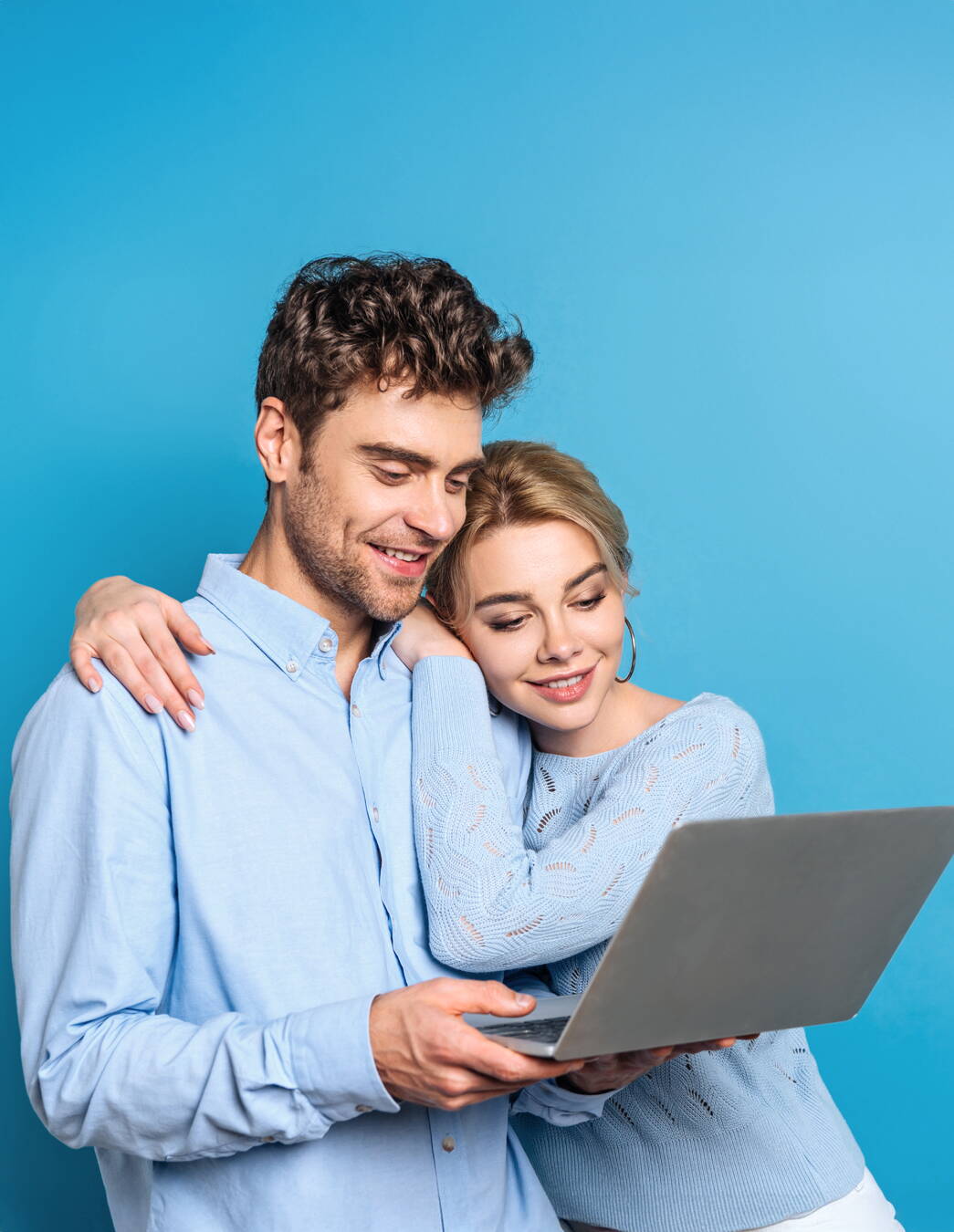 L’immagine mostra due persone che guardano insieme un computer portatile. Entrambe indossano abiti azzurri e si trovano davanti a uno sfondo blu. Una persona tiene il computer portatile, mentre l’altra le cinge le spalle con il braccio.